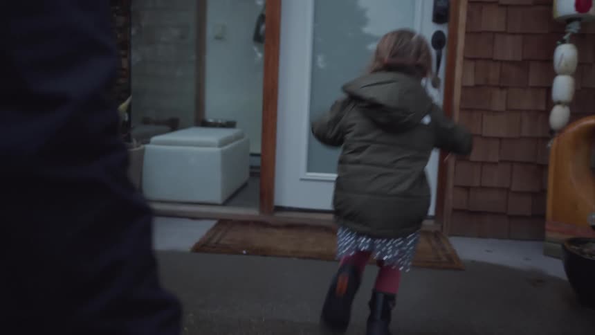 Father carries a suitcase while his young daughter moves forward to tug on the front door of a house on a winter day