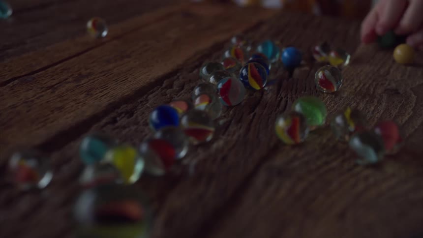A childs hand reaches for a scattering of colorful glass marbles spread across a rustic wooden surface