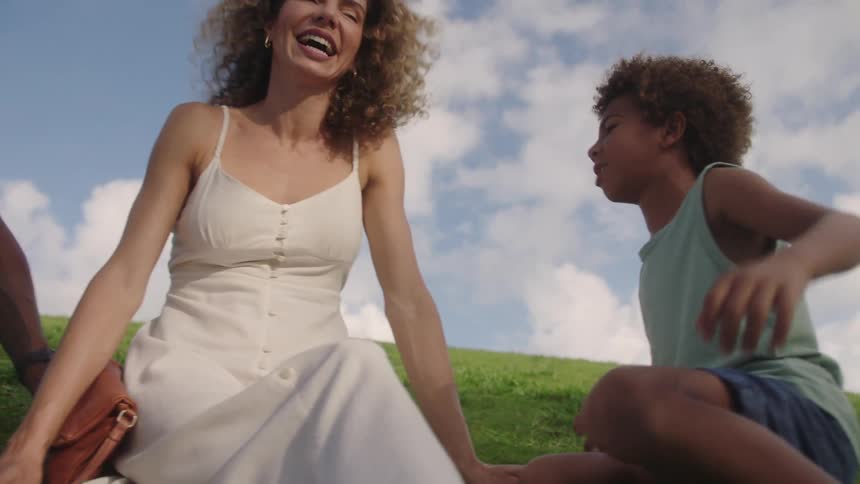 A family sitting on a grassy hill laughing under a blue sky with scattered white clouds