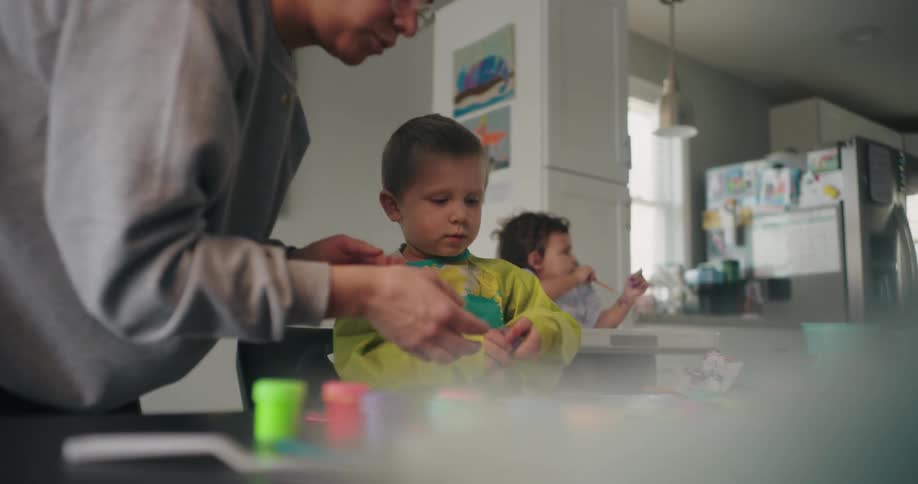 An adult is assisting a child with a craft activity at a table with colorful materials scattered across a home setting