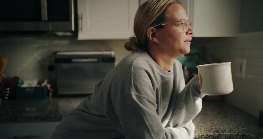 A woman holding a large ceramic cup reclines on the kitchen counter at home