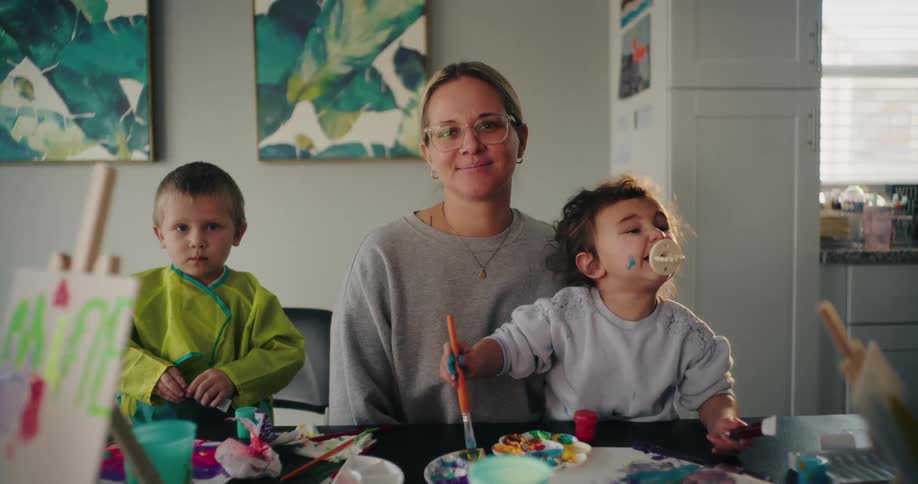 Mother with a happy facial expression holding a baby girl on her lap next to her other little son while they are playing with paint on a table