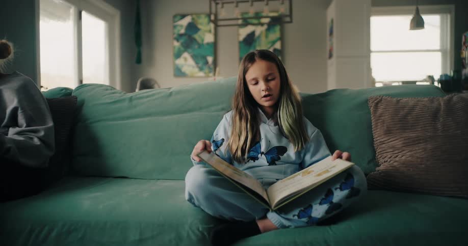 A girl in pajamas sits on a green couch in the living room at home reading a book