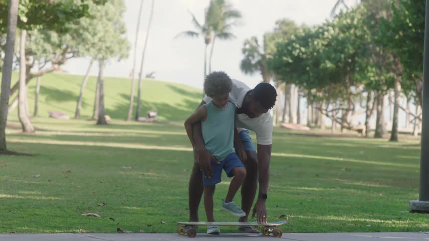 A father is teaching his son how to use a skateboard in a park