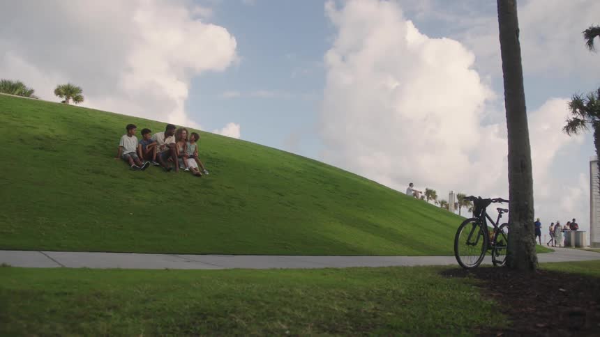 A family sits on a grassy hill in a park with a bicycle parked near a tree under a blue sky with scattered white clouds