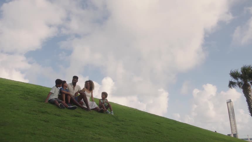 A family of five sitting on a lush green hill under a partly cloudy sky