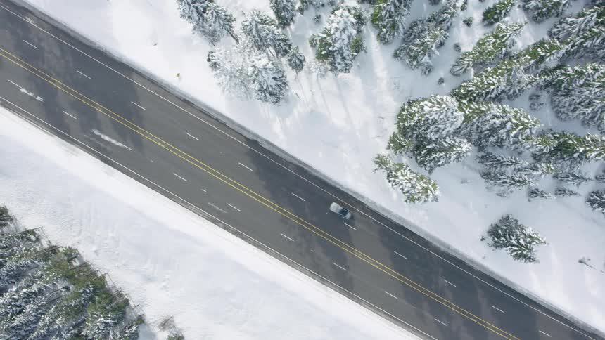 Birds eye view of a car driving on a plowed highway surrounded by sun kissed trees and snow