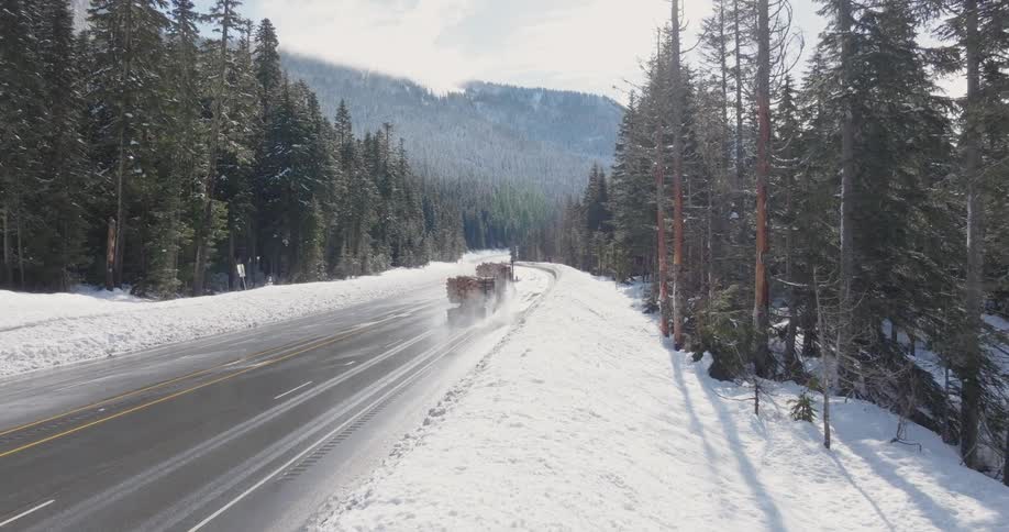 A logging truck creates a trail of mist and water spray as it heads up a snowy mountain pass highway
