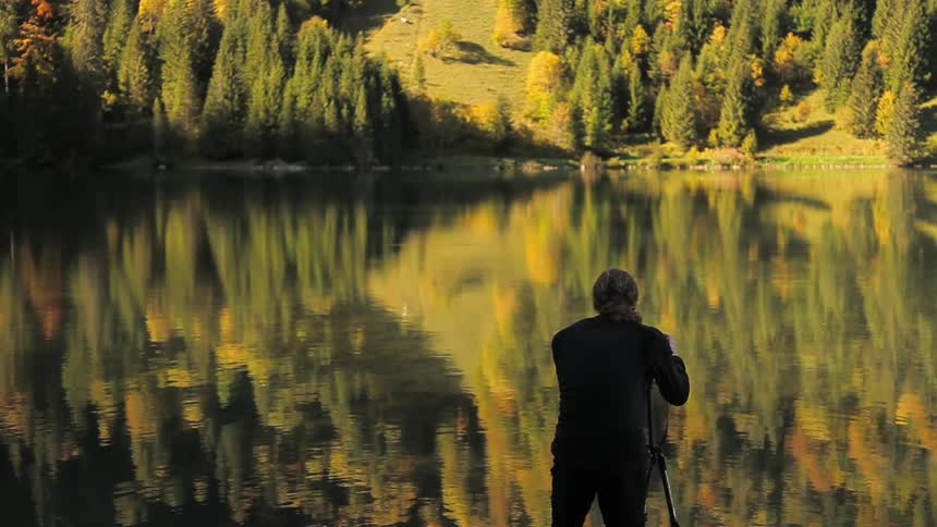 A man stands behind a tripod taking photos at a lake
