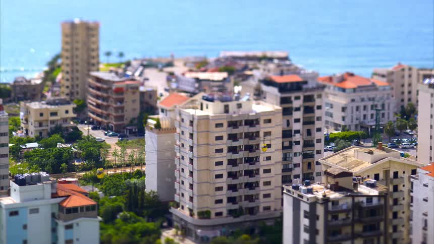 Seen from an elevated vantage with a tiltshift effect a coastal city features cable cars moving alongside buildings topped with orange roofs while the blue sea stretches into the distance beyond the clustered structures