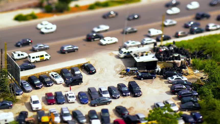A tilt shift aerial view of a parking lot next to a busy road