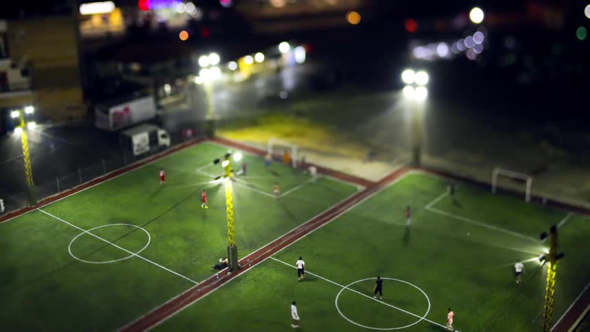 An aerial view shows a brightly lit outdoor soccer field at night with players engaged in a game