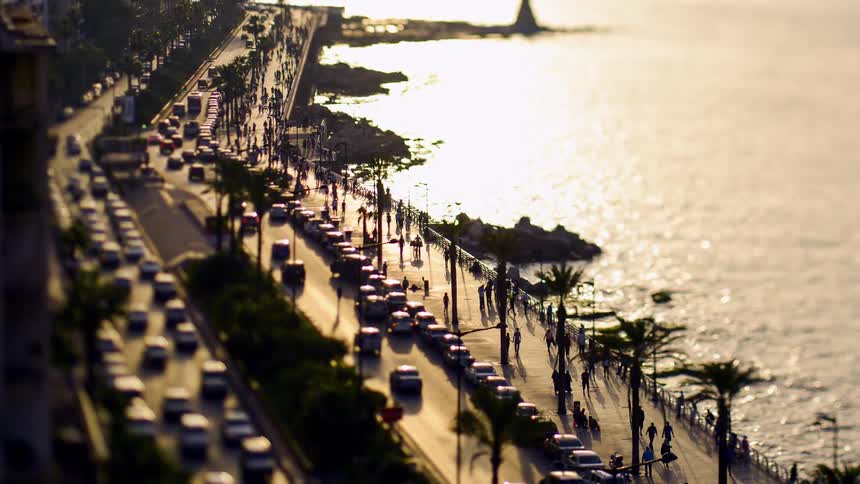 A bustling coastal highway lined with palm trees and cars running parallel to a shimmering body of water at sunset with people walking along the promenade