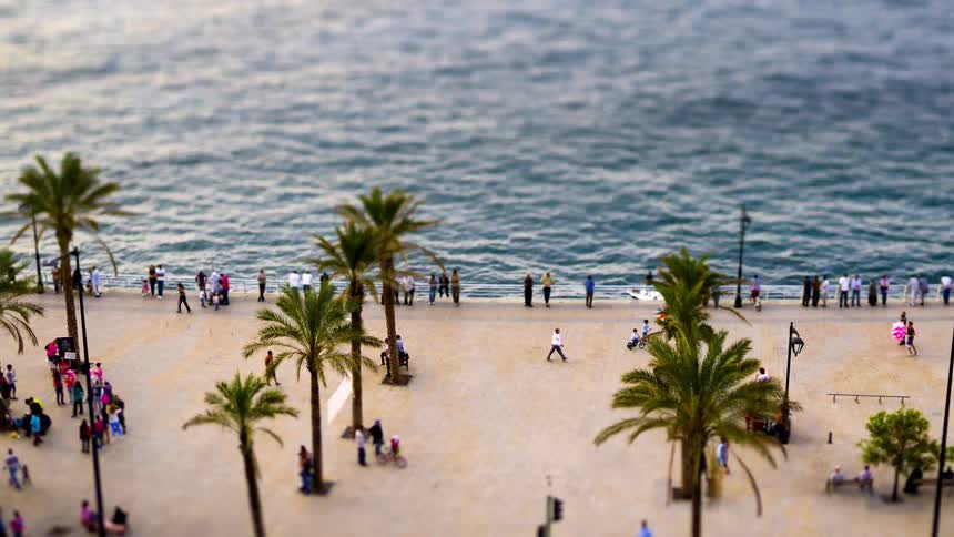 In a timelapse view people move along a coastal boardwalk as gentle waves lap the shore with a tiltshift filter lending a miniaturelike effect to the bustling seaside scene beneath swaying palm trees