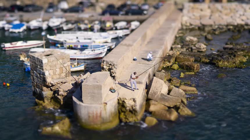 In a tiltshift timelapse people fish from a seaside concrete pier while boats moored in a small marina and floating buoys accent the waters edge creating a miniaturelike view of the bustling waterfront
