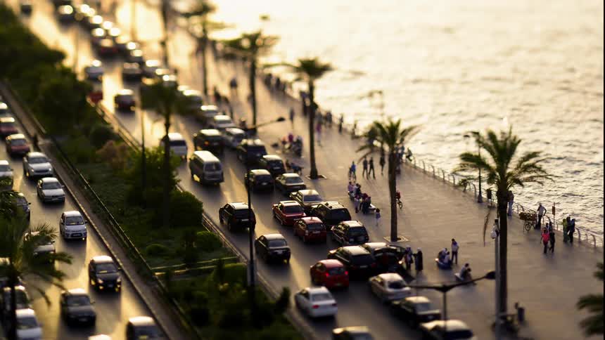 There are groups of people on the sidewalk beside of the water and there are lots and lots of cars parked along the road and driving on the two sections of road that are going different directions