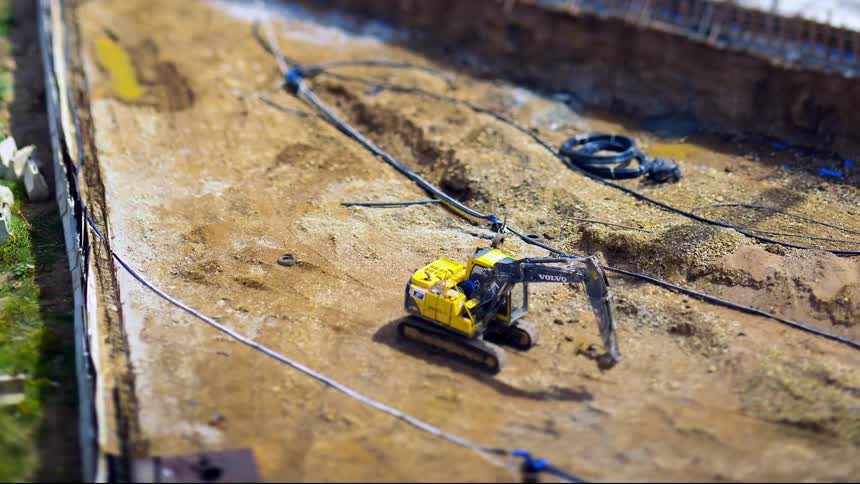 From a high vantage with a tiltshift effect a person enters an excavator and drives out of frame across the dusty site while other construction workers walk away highlighting a busy scene of ongoing work