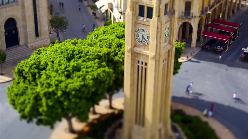 A beige clock tower with two visible clock faces set among vibrant green trees and a lively plaza with people and vehicles
