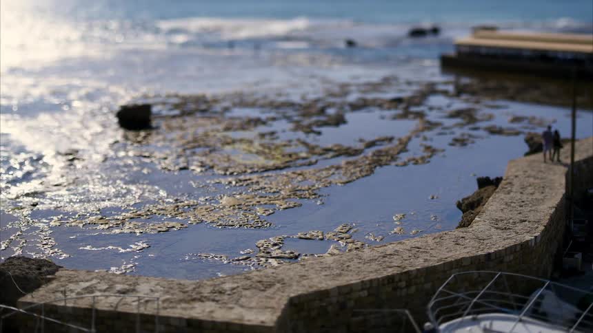 The man and woman are walking down the saidewalk that is next to the water that has slime and vegetation on the top of it