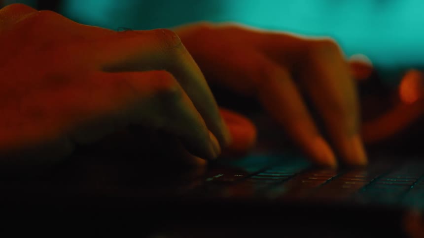 Womans hands typing on a laptop keyboard illuminated by warm orange light against a dark background