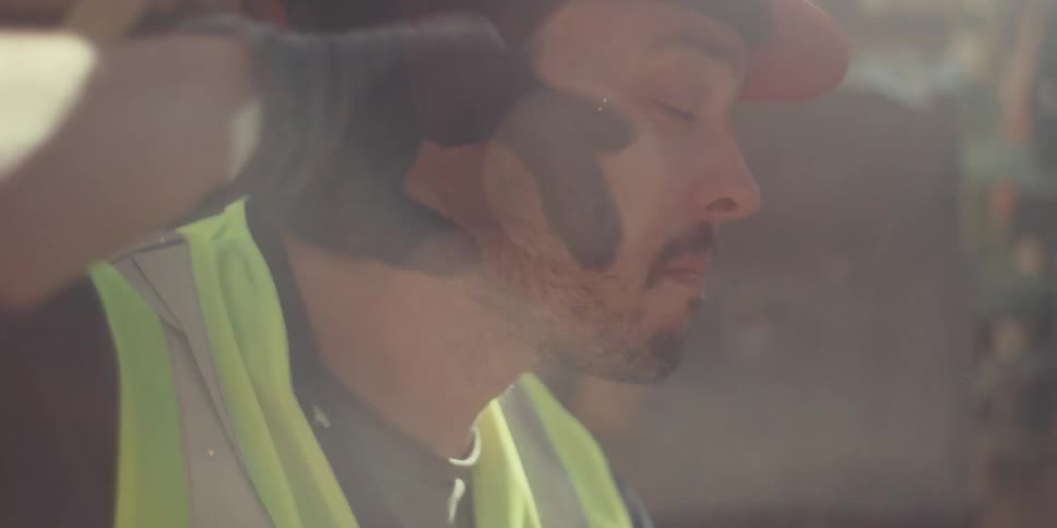 A construction worker wearing a hard hat and gloves wipes up the sweat from his eyes