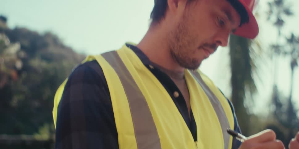 A construction worker wearing a vest and hardhat takes notes on a clipboard under the bright sunlight of a clear day