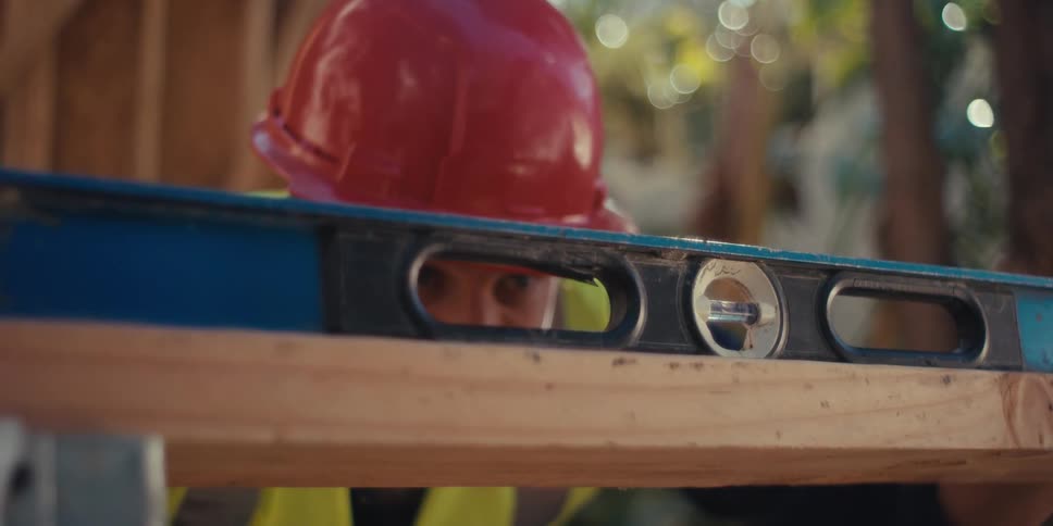 A construction worker wearing a vest and a hardhat places a level over a plank of wood