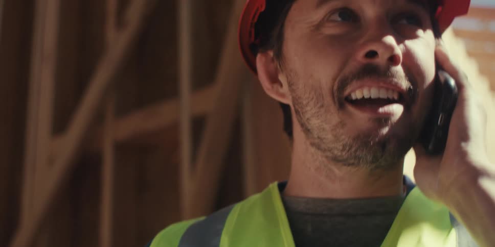 A construction worker in a safety vest and hard hat talks on the phone while inspecting a clipboard