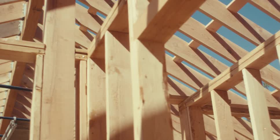Sunlight illuminates the wooden framework of a building under construction revealing angled beams and a bright blue sky through the gaps