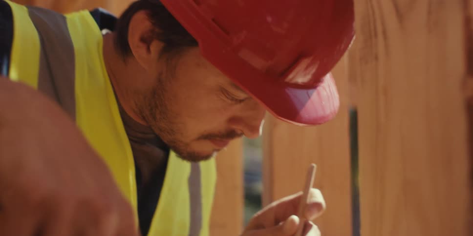 A construction worker wearing a vest and hardhat measures a planks length with a tape measure and marks it with a pencil