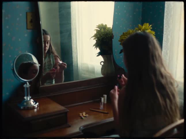 Girl applying lip gloss while sitting in front of a mirror in a room