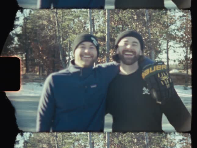 Two smiling men in winter hats and hockey gloves pose for a photo on an ice rink surrounded by trees