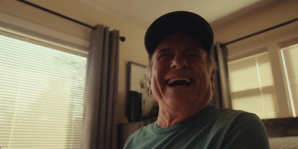 Elderly man with cap and blue tshirt uses tablet laughing in a room