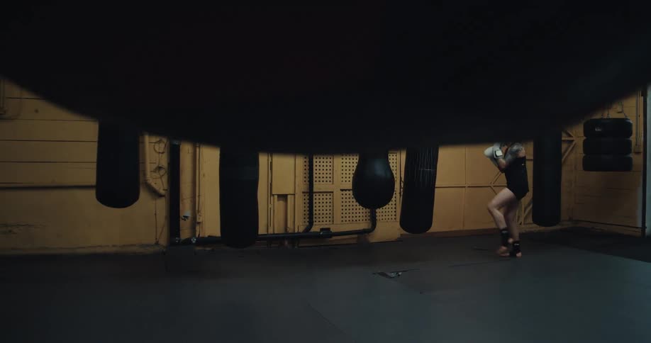 A strong woman with tattooed arms and braided hair practices punching a bag in a gym while wearing white boxing gloves