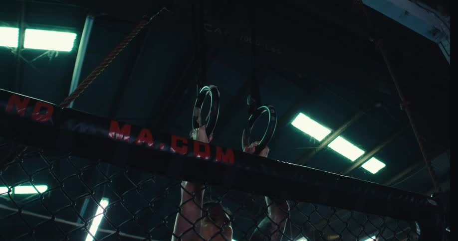 A strong tattooed woman lifts herself on gymnastic rings performing an exercise in a dimly lit gym