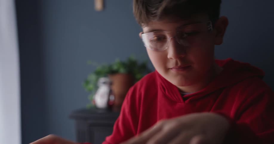A boy in a red sweater proudly admires an object he assembled on a desk in his room