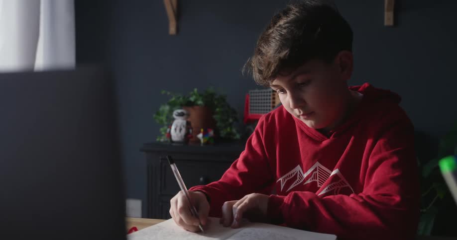 A young boy in a red hoodie is sitting at a wooden desk writing in a notebook with a focused expression while a laptop and a cup of pens are nearby in a dimly lit room
