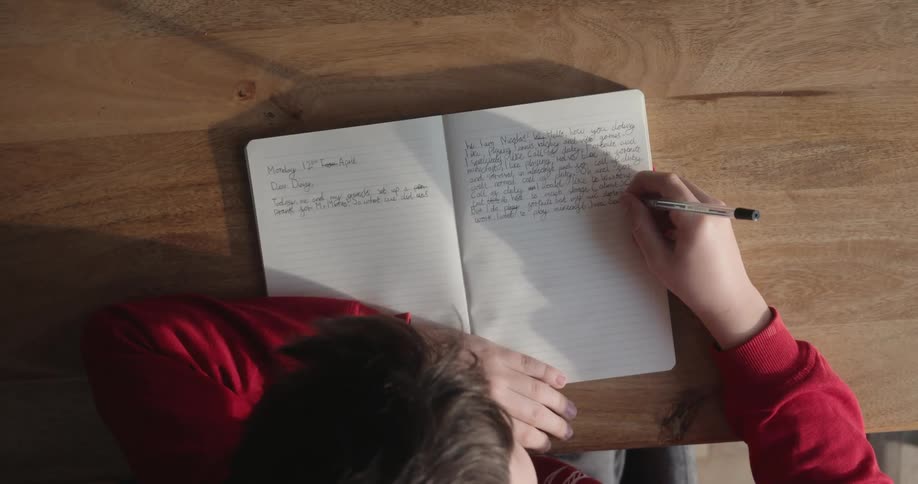 Overhead view of young boy sitting at wood table writing in a journal