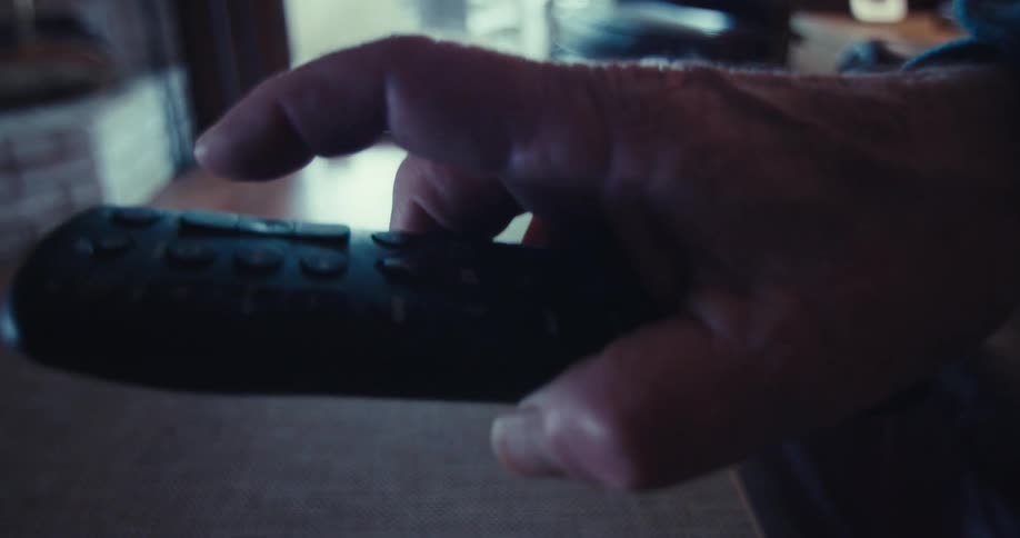A hand reaches for and uses a remote control lying on a table in a dimly lit living room