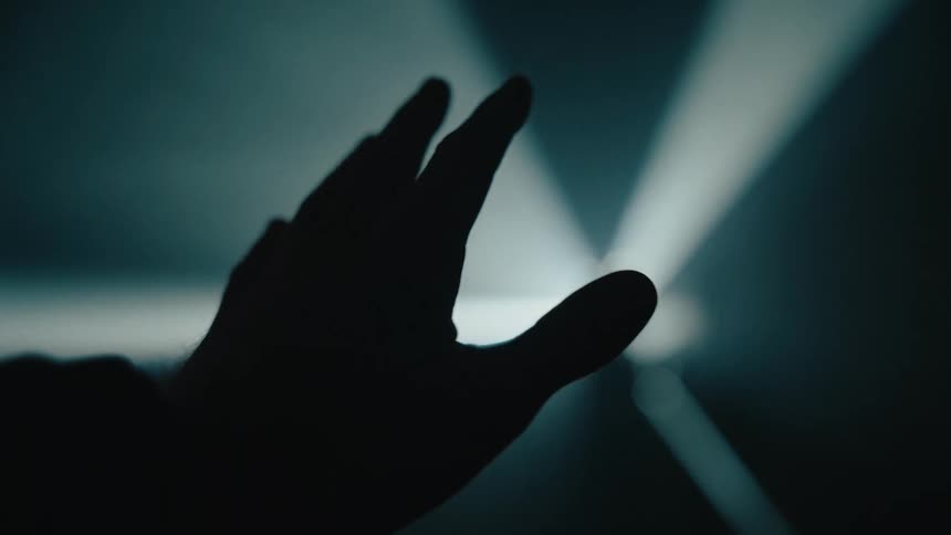 A silhouetted hand reaches into a dimly lit space its fingertips faintly illuminated by a soft glow