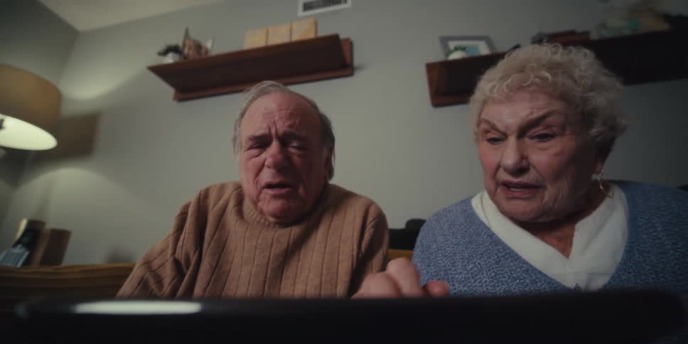An elderly couple sits together appearing to react with concern or frustration while looking at something on a screen