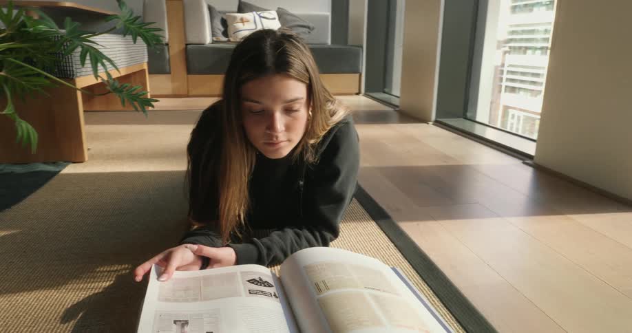 A teen girl is reading a book while laying on the floor near a window