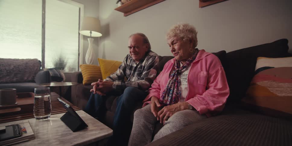 Two grandparents are sitting on the couch while talking to their grandchildren on a tablet