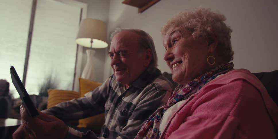 Elderly couple sitting in their living room smiles while sharing a tablet during a video call