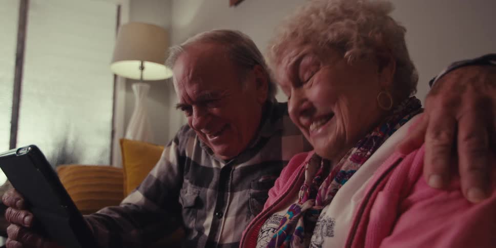 An elderly couple sits on the couch laughing at something theyre watching on a tablet