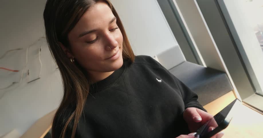 A woman with straight hair wearing a dark sweatshirt and light colored pants is using her smartphone to text in the living room of an apartment