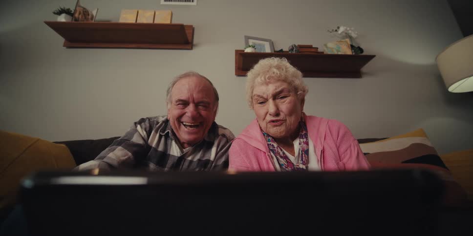 Elderly couple sitting in their living room smiles and waves during a video call