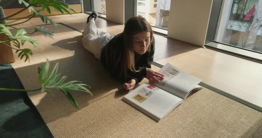 A person reclines on a mat in a sunlit room reading an open book amidst potted plants and large windows