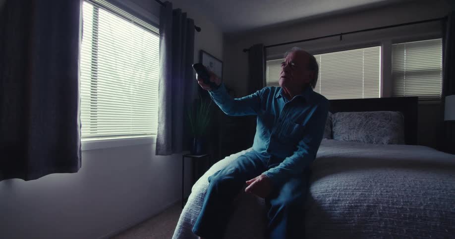 Elderly man sitting on his bed gestures and talks to himself while using his television remote