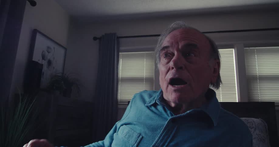 An elderly man sits in a dimly lit room holding a remote control as he turns on the tv appearing surprised before bursting into laughter